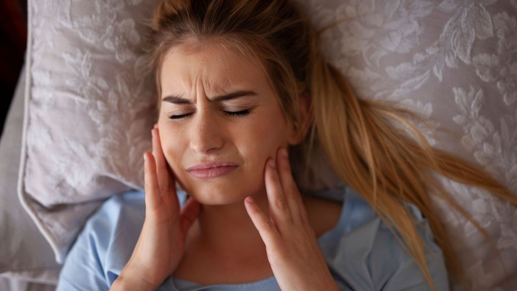 Woman in bed holding her jaw in pain from teeth grinding
